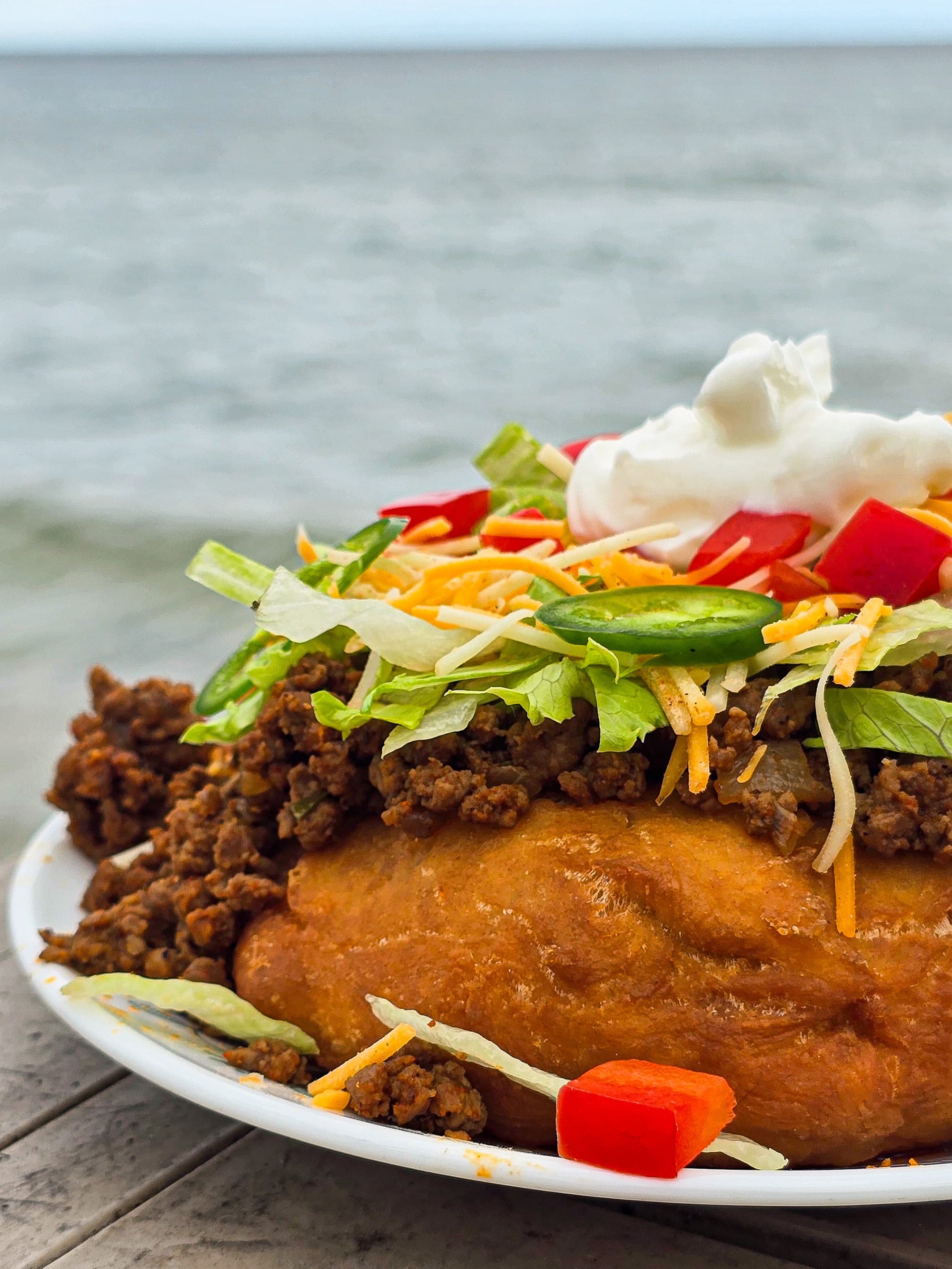 An Indian Taco made from a piece of frybread, seasoned ground meat taco filling, lettuce, chopped red peppers, cheese, sour cream, and jalapeno pepper slices.
