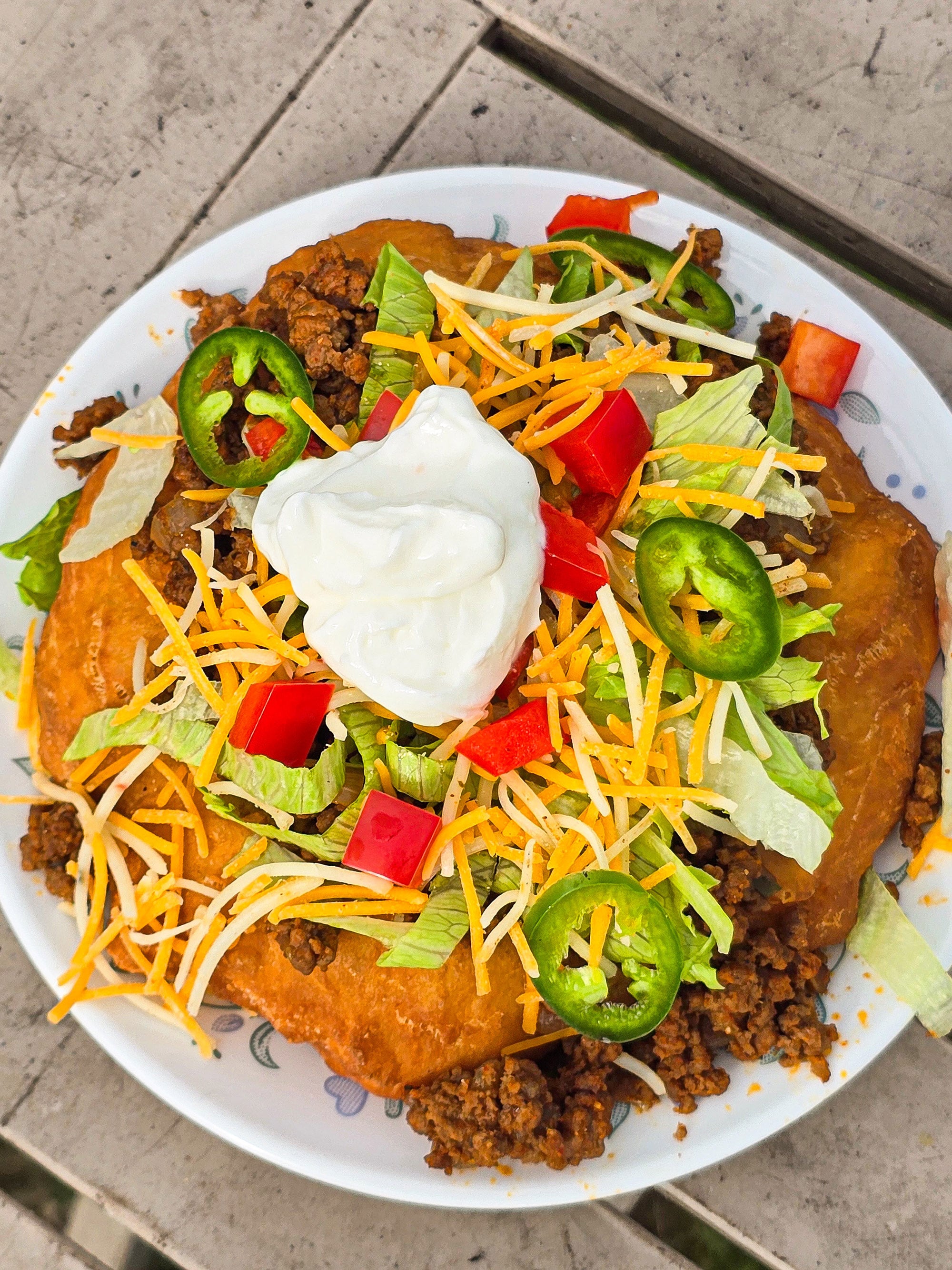 And Indian Taco made from a piece of fry bread, seasoned ground meat taco filling, lettuce, chopped red peppers, cheese, sour cream, and jalapeno pepper slices.