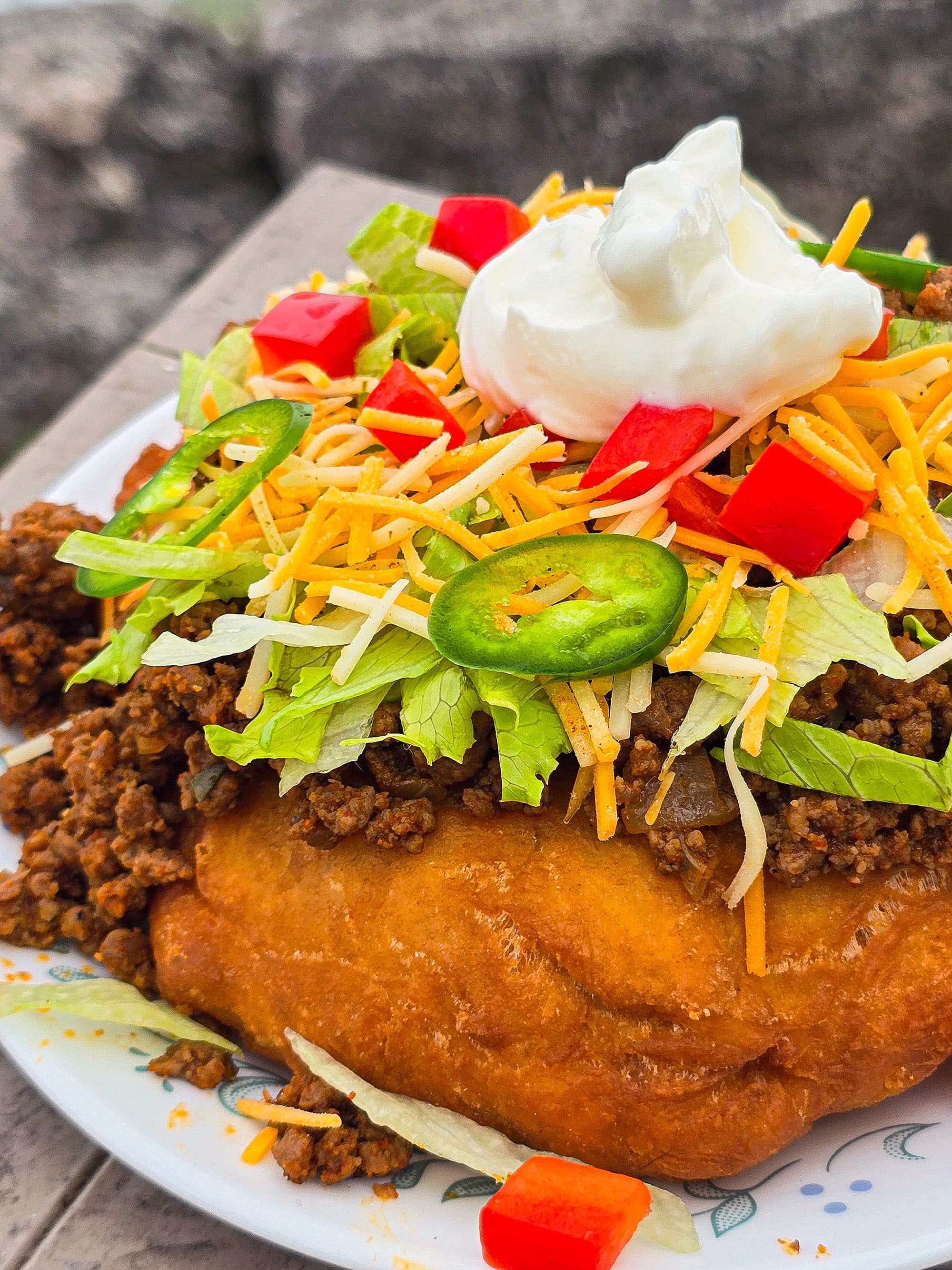 An Indian Taco made from a piece of frybread, seasoned ground meat taco filling, lettuce, chopped red peppers, cheese, sour cream, and jalapeno pepper slices.