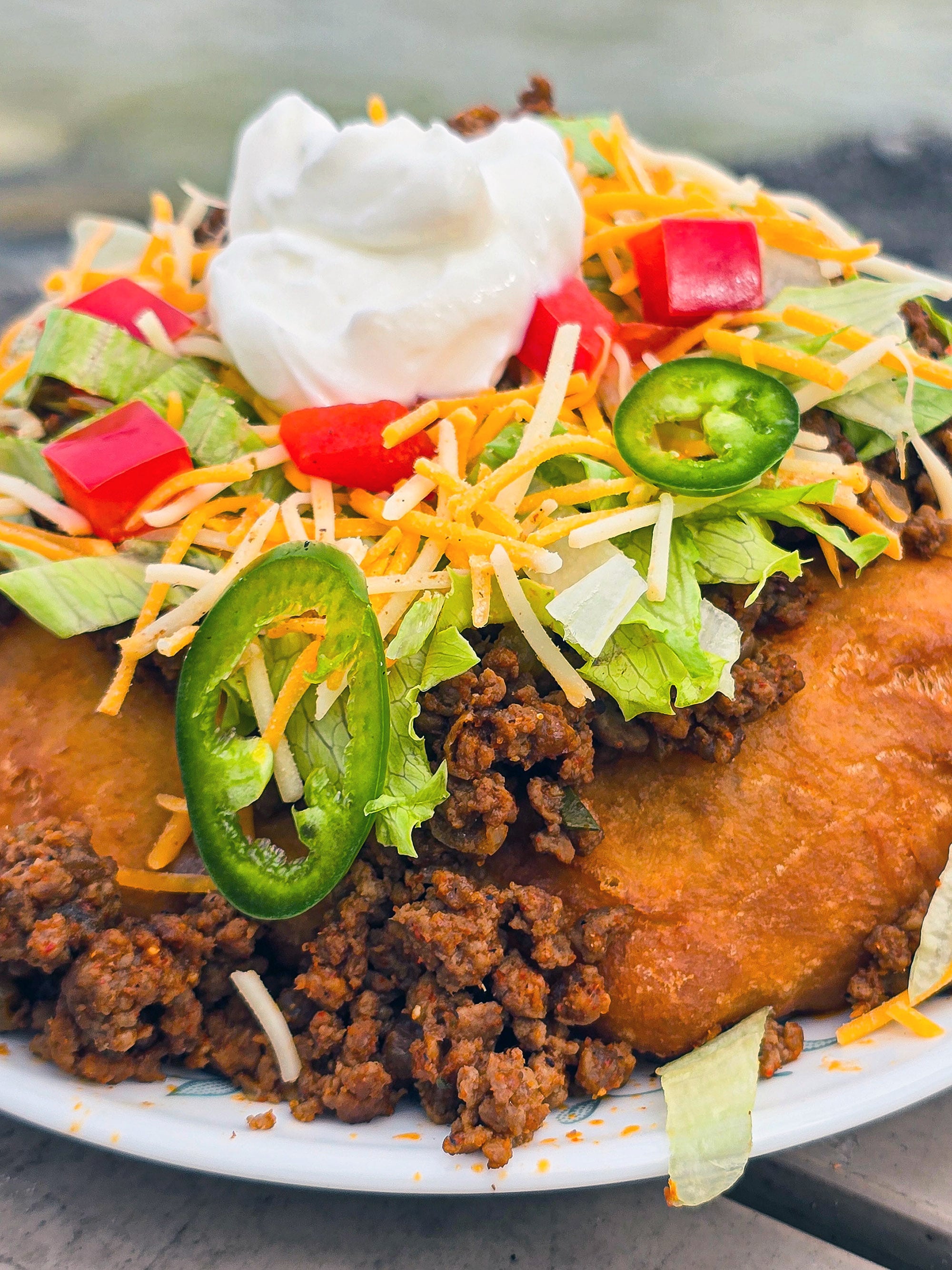 And Indian Taco made from a piece of fry bread, seasoned ground meat taco filling, lettuce, chopped red peppers, cheese, sour cream, and jalapeno pepper slices.