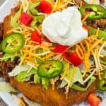 An Indian Taco made from a piece of frybread, seasoned ground meat taco filling, lettice, chopped red peppers, cheese, sour cream, and jalapeno pepper slices.