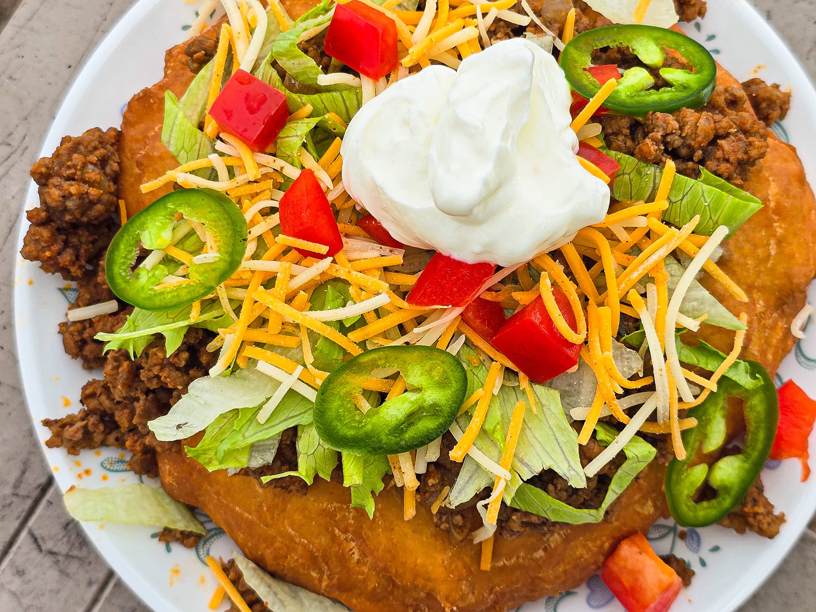 An Indian Taco made from a piece of frybread, seasoned ground meat taco filling, lettuce, chopped red peppers, cheese, sour cream, and jalapeno pepper slices.