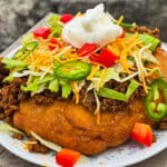 And Indian Taco made from a piece of fry bread, seasoned ground meat taco filling, lettice, chopped red peppers, cheese, sour cream, and jalapeno pepper slices.