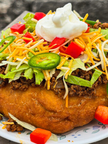 And Indian Taco made from a piece of fry bread, seasoned ground meat taco filling, lettice, chopped red peppers, cheese, sour cream, and jalapeno pepper slices.