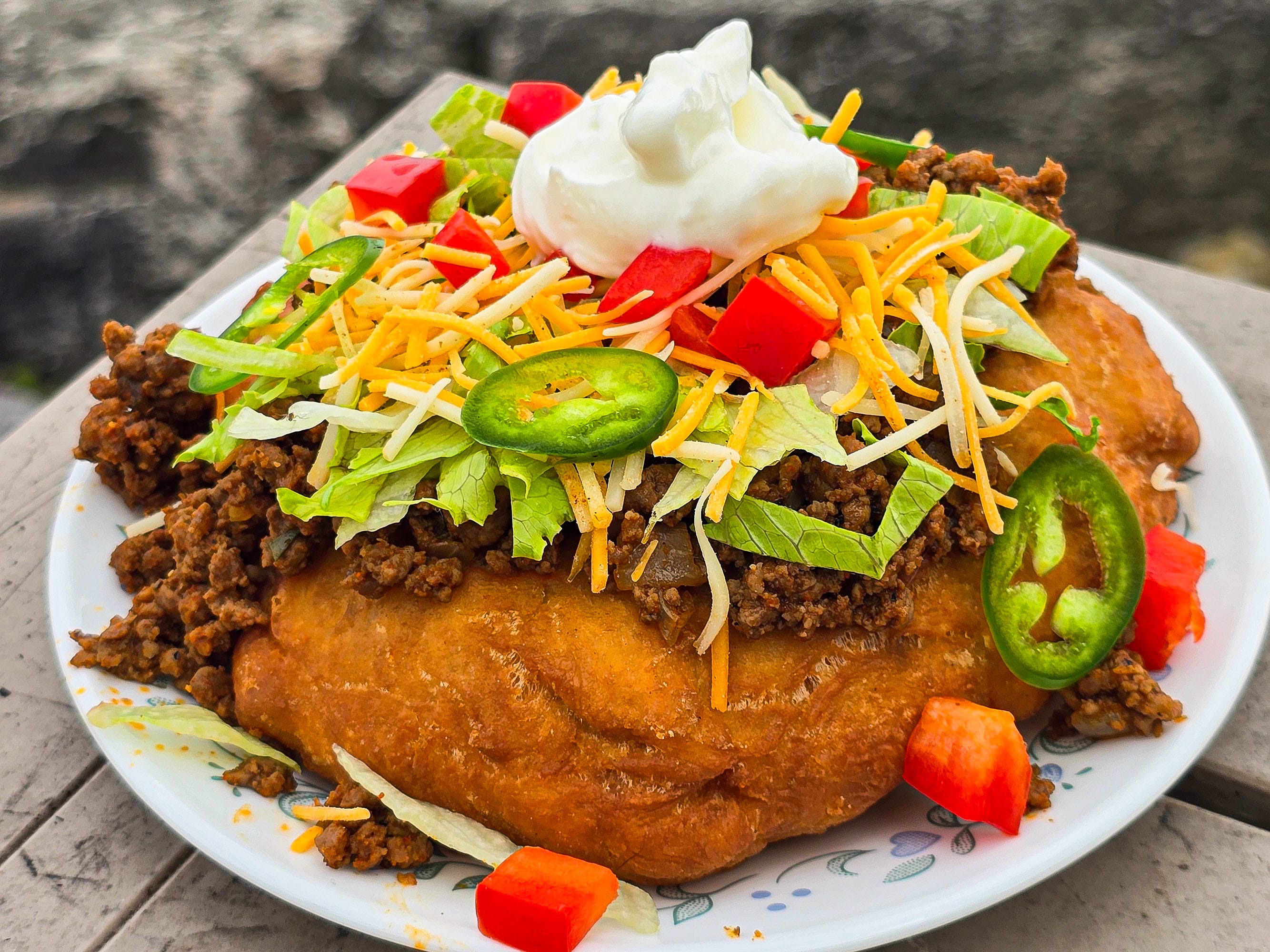 And Indian Taco made from a piece of fry bread, seasoned ground meat taco filling, lettuce, chopped red peppers, cheese, sour cream, and jalapeno pepper slices.