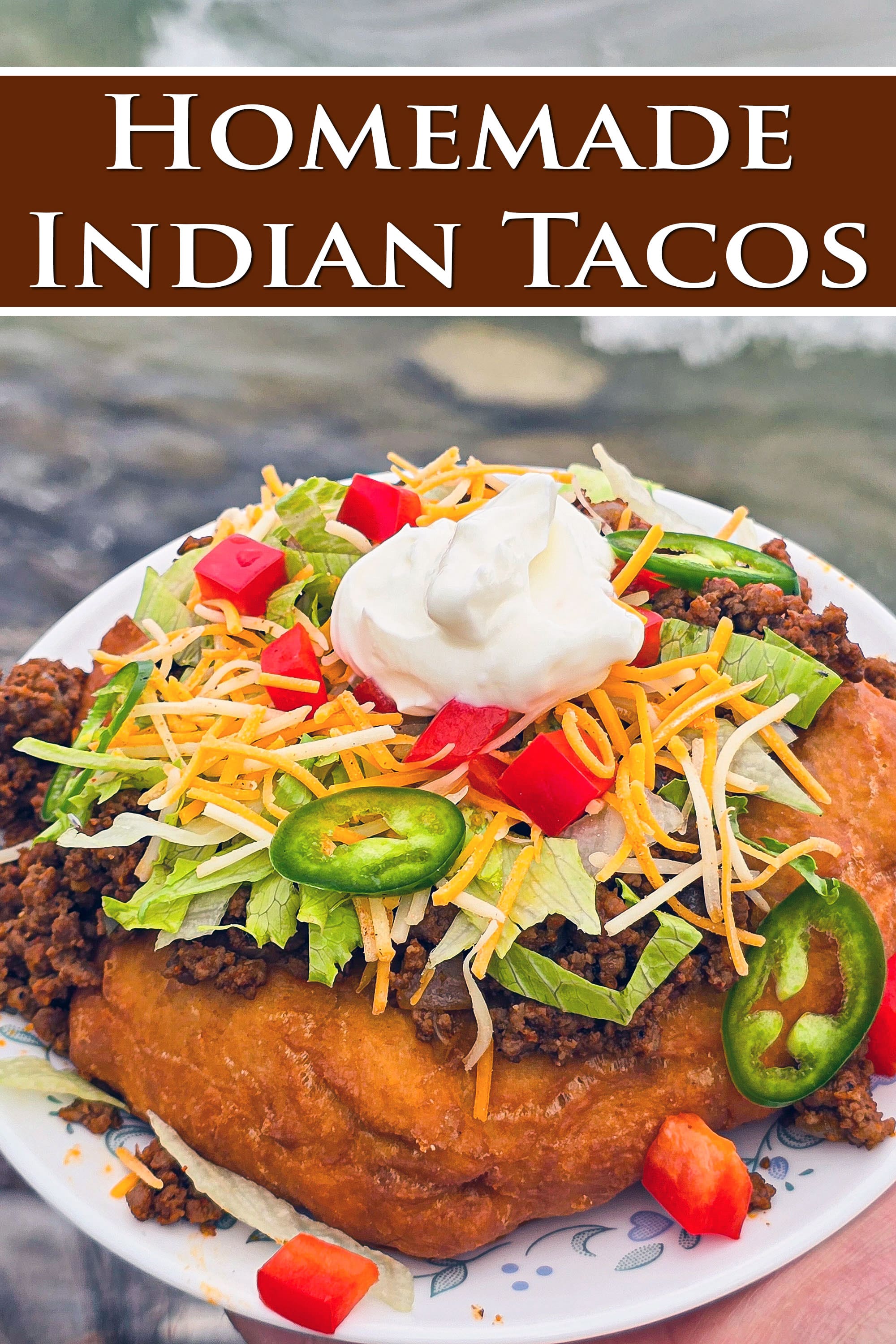 An Indian Taco made from a piece of frybread, seasoned ground meat taco filling, lettuce, chopped red peppers, cheese, sour cream, and jalapeno pepper slices. Overlaid text says homemade Indian tacos.