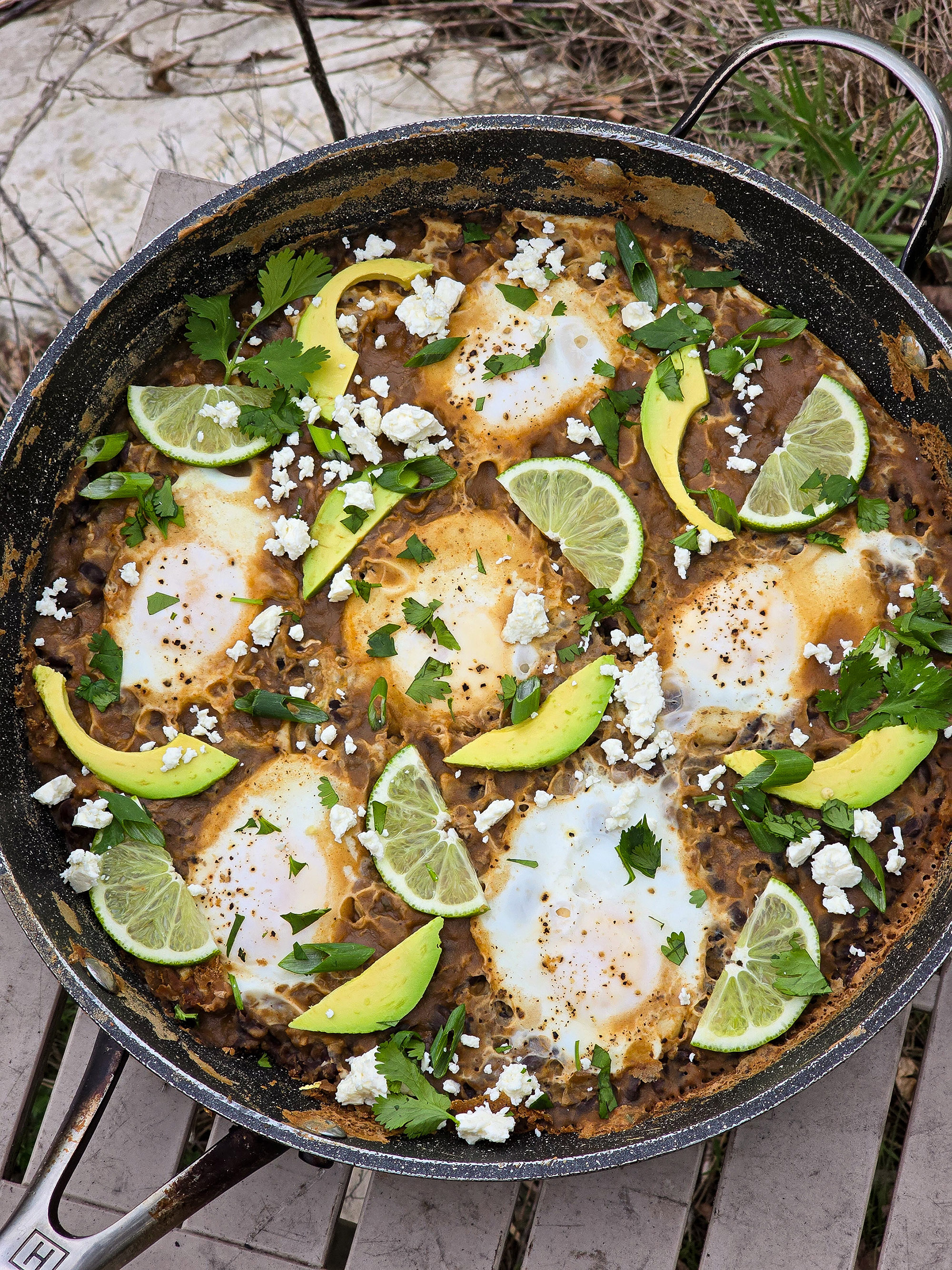 A pan of eggs in spicy black beans, garnished with lime slices, avocado, cilantro, and crumbled queso fresco.