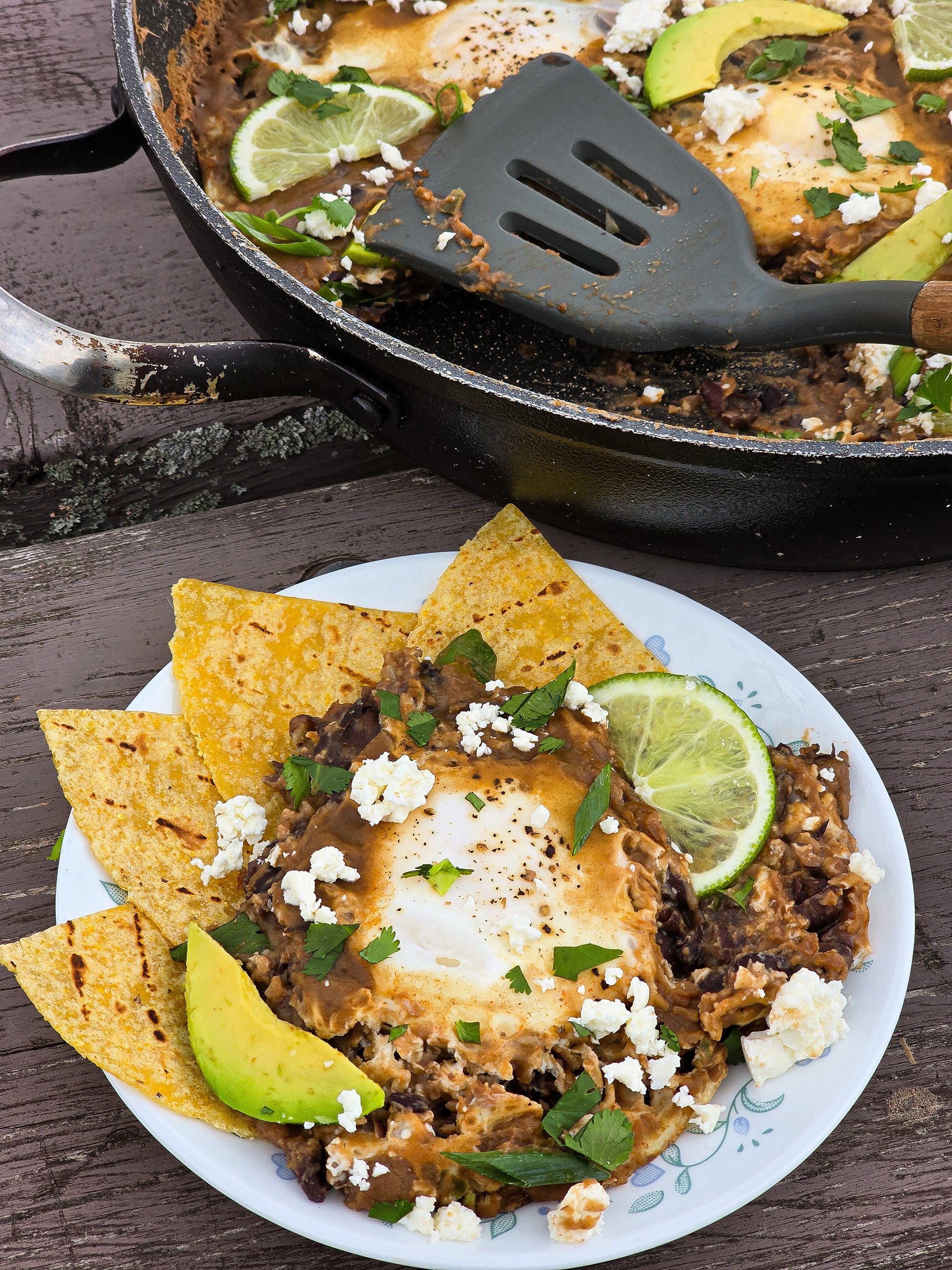A pan of Huevos Enfrijolados, garnished with lime slices, avocado, cilantro, and crumbled queso fresco.