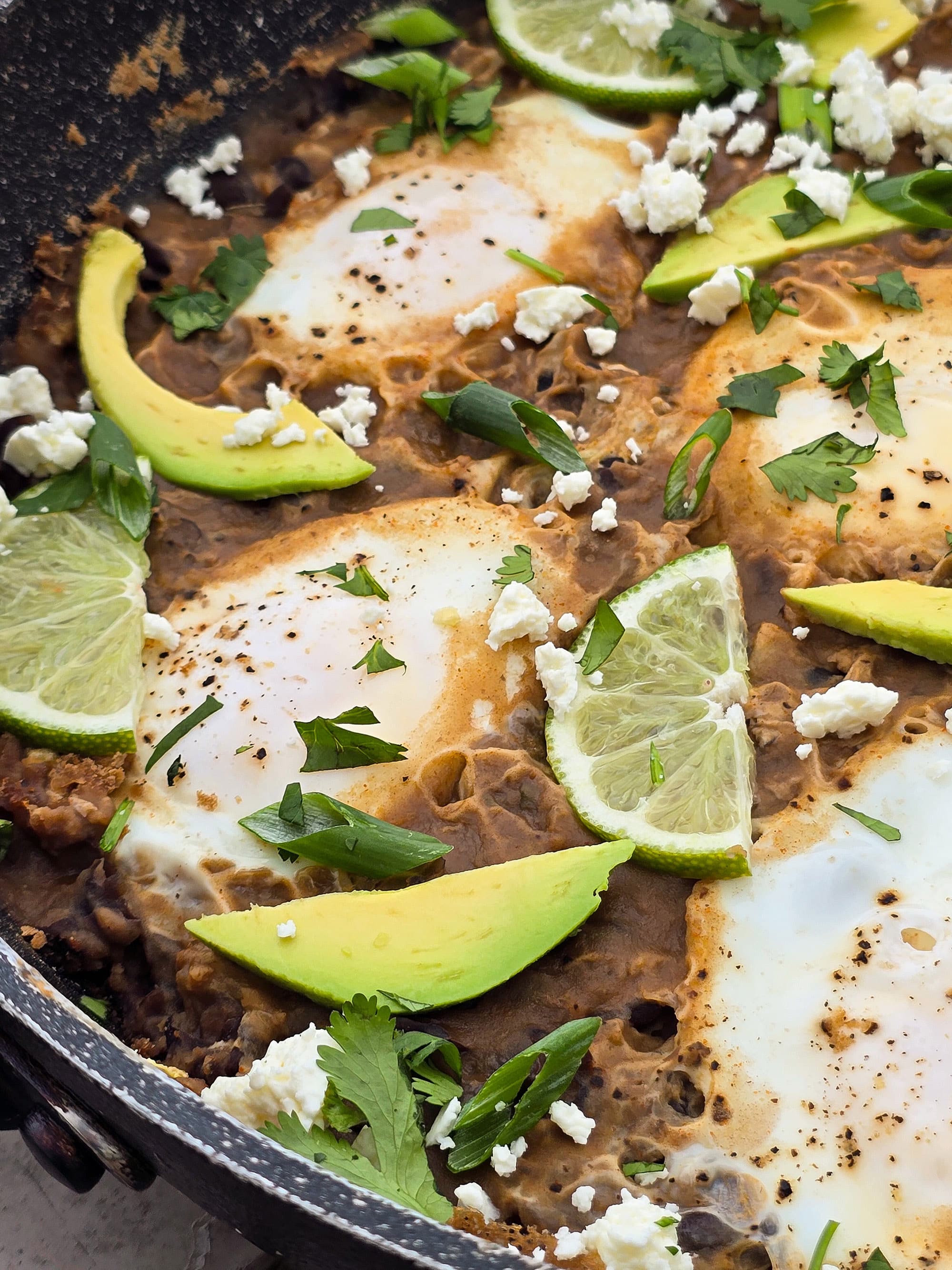 A pan of eggs in spicy black beans, garnished with lime slices, avocado, cilantro, and crumbled queso fresco.