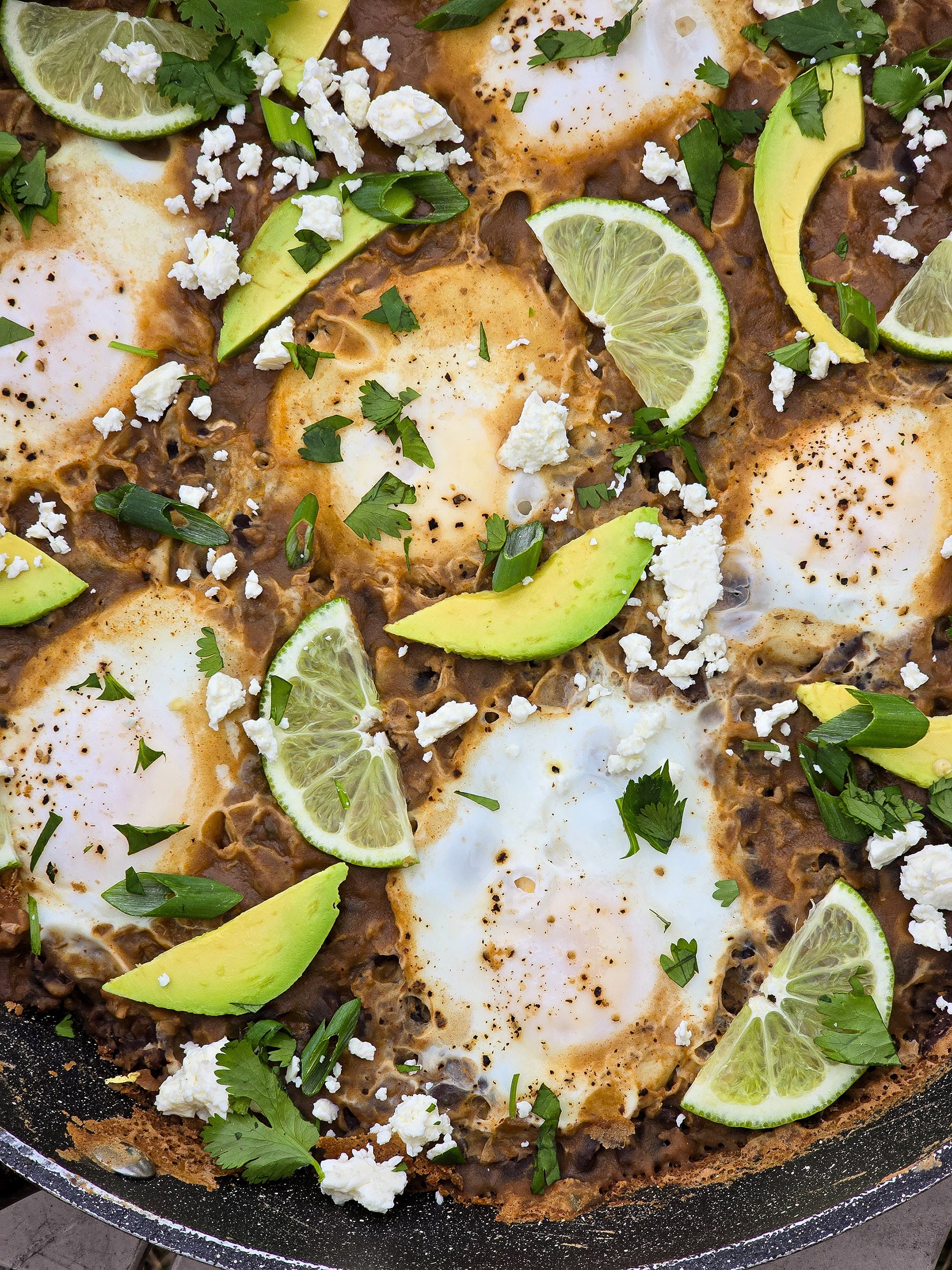 A pan of Huevos Enfrijolados, garnished with lime slices, avocado, cilantro, and crumbled queso fresco.