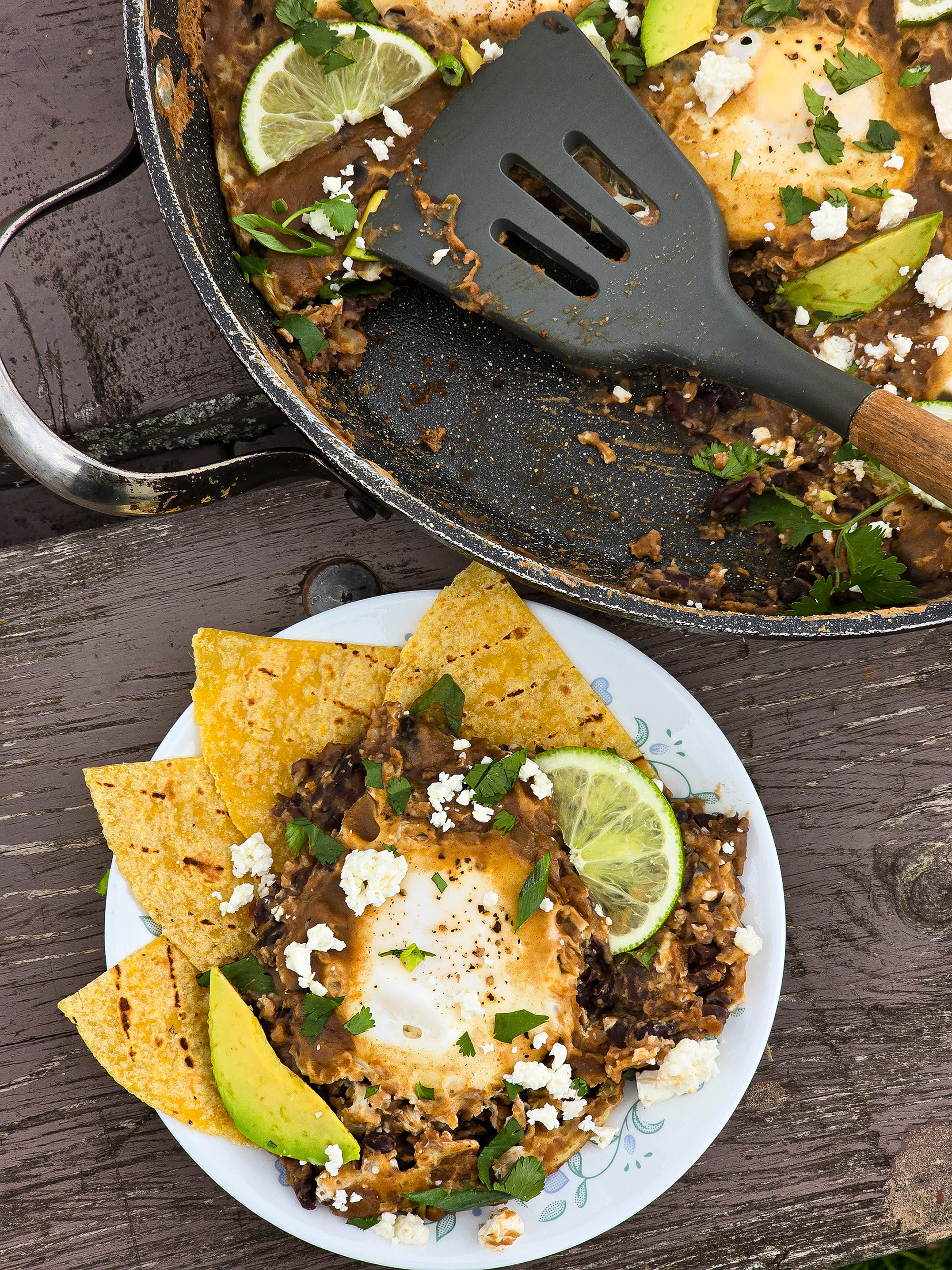 A pan of eggs in spicy black beans, garnished with lime slices, avocado, cilantro, and crumbled queso fresco.