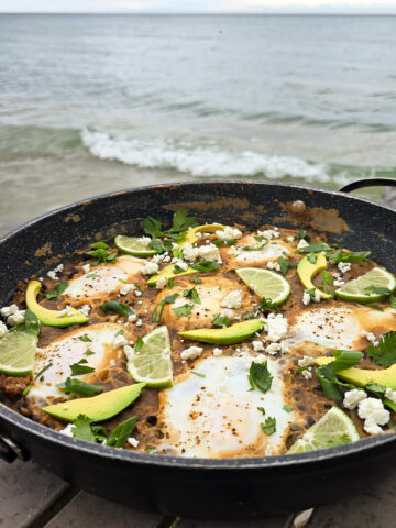 A pan of Huevos Enfrijolados, garnished with lime slices, avocado, cilantro, and crumbled queso fresco.