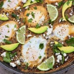 A pan of eggs in spicy black beans, garnished with lime slices, avocado, cilantro, and crumbled queso fresco.