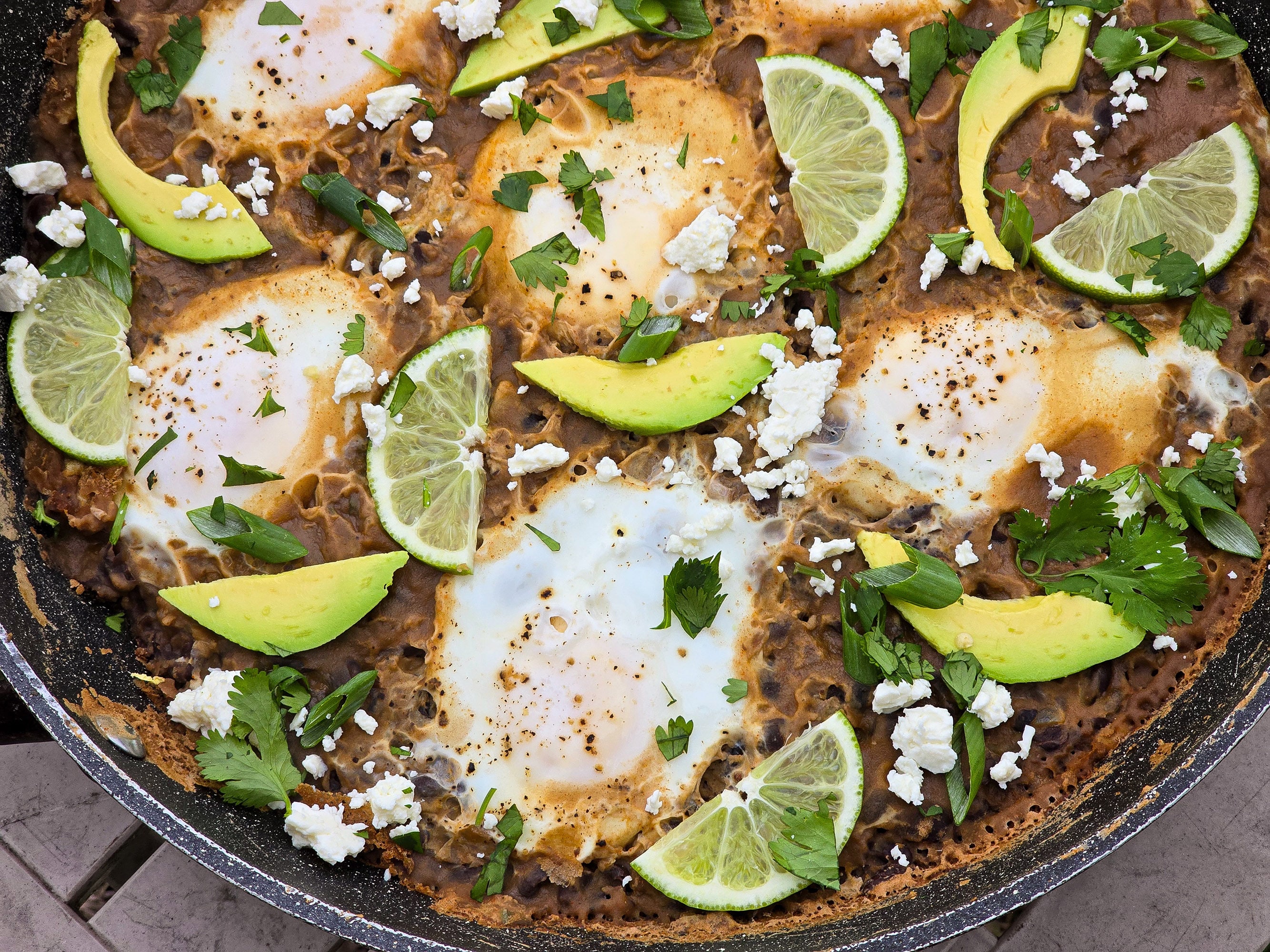 A pan of eggs in spicy black beans, garnished with lime slices, avocado, cilantro, and crumbled queso fresco.